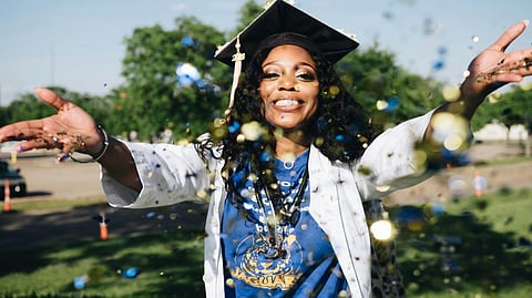 A graduate wearing a white coat, a graduation cap, a blue t shirt is throwing confetti.
