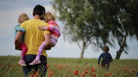 A father carries two girls in a field while a boy runs ahead, enjoying a sunny day outdoors.