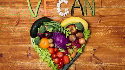 Vegetables arranged in the shape of a heart on a wooden table and the word 'VEGAN' spelled out.