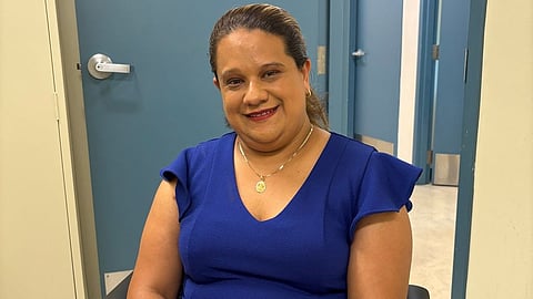 A woman sits in a chair, wearing a blue dress with flutter sleeves, in a brightly lit room.
