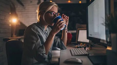 A person seated at a desk, holding a blue cup, working intently on a computer at night.