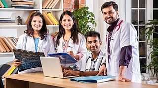 A group of four medical professionals in lab coats collaborates at a desk with a laptop, medical files, and an X-ray.