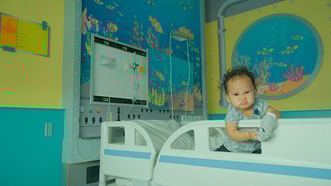 A toddler sitting in a hospital bed for children with colorful paintings in the background.
