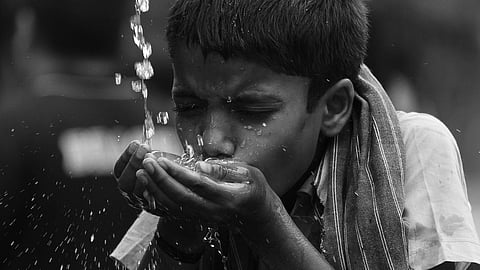 A young boy drinks water from a flowing tap, using his hands. Wet hair and shirt suggest relief from thirst.