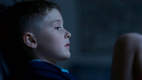 A young boy sitting in a dimly lit room, lost in deep thought, reflecting emotional stress.