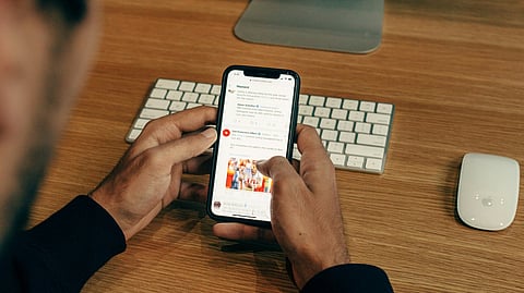 A man is sitting in front of his laptop, checking news updates on his mobile phone.