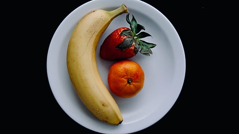 A white plate with a banana, strawberry and an orange.