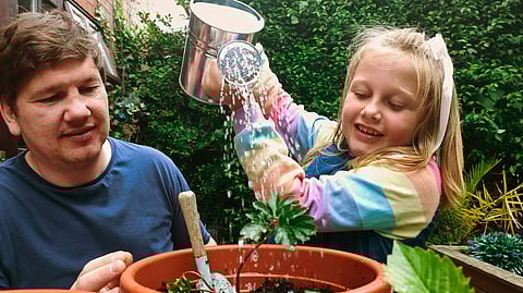 A young girl waters a potted plant while smiling, as her father looks on supportively. 