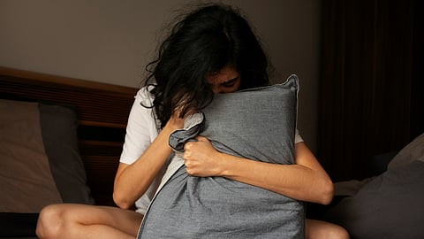 Shows woman sitting on a bed holding a pillow in distress due to poor sleep.