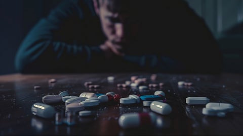 A man is seated at a table with numerous pills strewn around him, suggesting a moment of distress or contemplation.