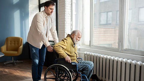 An elderly man in a wheelchair is being helped up by a young male near a window indoors.