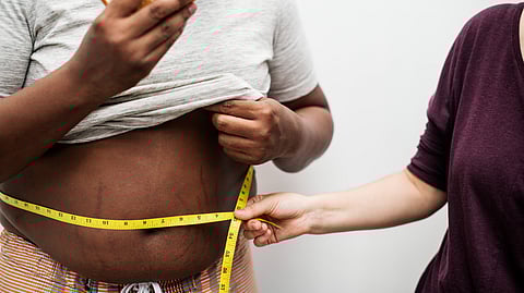 A person measuring an obese man's waist while he is eating a burger.