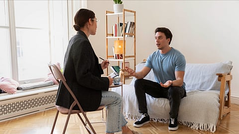 A young man sitting on a couch and talking to a lady wearing a black blazer.