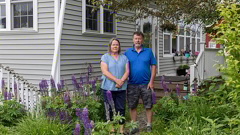 A man and woman stand together in front of their home, smiling and posing for the camera.