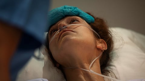 A woman lies in a hospital bed wearing a nasal oxygen tube, looking up with a concerned expression. She has a blue medical cap resting on her forehead and is being attended to by a healthcare worker.