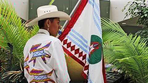 A man wearing a cowboy hat and standing with holding a flag of his tribe.