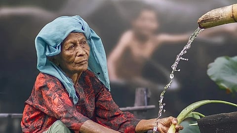 An elderly woman, looking sad, fills water using a bamboo container.