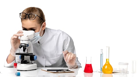 A woman is observing something in a microscope and other lab equipment like pippettes, beakers nearby.