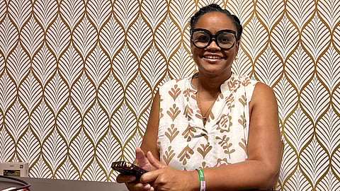 Image depicting smiling woman holding a phone, seated against a patterned wallpaper.