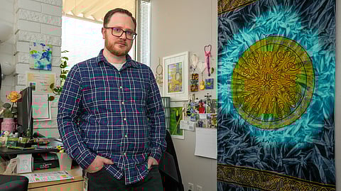A man in a blue plaid shirt stands casually in a decorated office space, next to a wall hanging with an intricate circular design in vibrant green, yellow, and blue tones.
