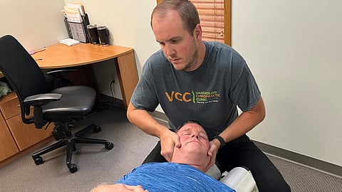 A man lies on a chiropractic table while the chiropractor adjusts his leg. They are in a clean clinic room with posters and equipment.