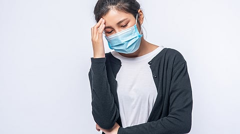 Image depicting woman wearing a face mask holding her head, appearing unwell.