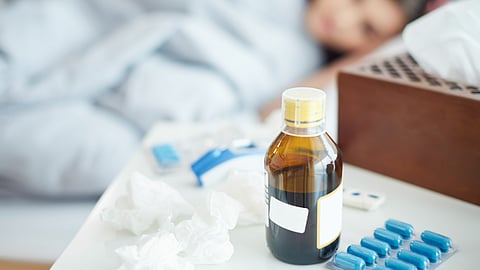 Close-up of a medicine bottle, blue capsules, and tissues on a bedside table, with a sick person lying in bed in the background.