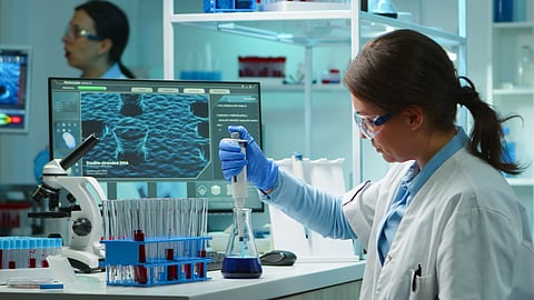 A scientist in a lab coat and gloves uses a pipette to handle a blue liquid in a flask, surrounded by test tubes, a microscope, and a computer displaying molecular analysis data, with another researcher working in the background.