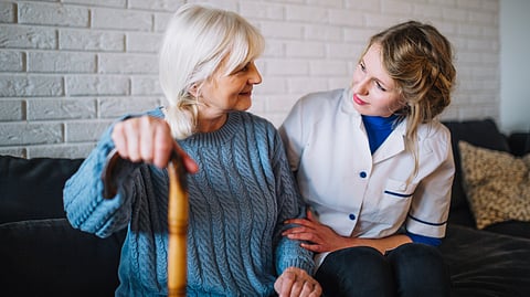 An elderly lady talking to a nurse sitting on a couch clutching a walking stick.