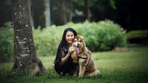 A smiling woman with long dark hair embracing a fluffy dog with brown and white fur, in a natural, outdoor setting.