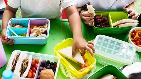 Image shows children eating from lunchboxes filled with sandwiches, fruits and snacks.