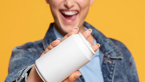 Image of a woman smiling and showing a can of energy drink standing in an orange background.