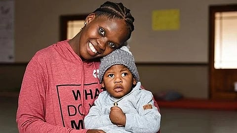 A black woman wearing a pink top holding a small boy in grey hoodie in her lap indoors.