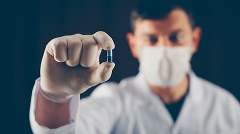 A doctor with mask and gloves holding blue pill with his fingers.