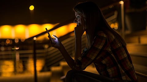 A young woman sitting in dark, outside on stairs looking at her phone.