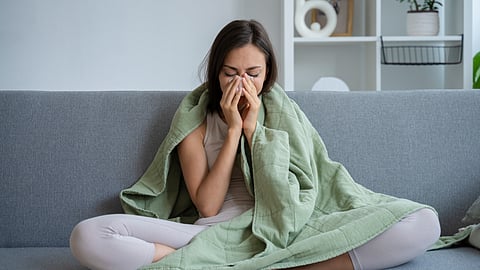 A woman sitting on a couch, covered with a blanket, pinching her nose as she is sick.