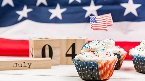 A July 4 theme cupcake and American flag in the background with July 4 written with wooden tiles on the side.