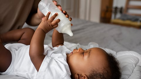 A baby is holding a milk bottle and looking at it while lying down on a bed.