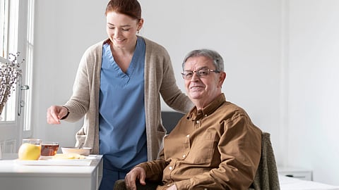 Smiling elderly man sitting with a caregiver providing support and preparing a meal in a clinical setting.