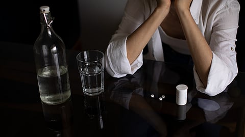 A woman holds her face in her hands at a table with drinks, pills and glasses. 
