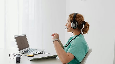 A healthcare professional in scrubs sits at a desk, using a laptop and wearing a headset. Sunlight filters through curtains, creating a calm atmosphere.