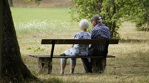 An old couple sitting on a wooden bench in a park outside.