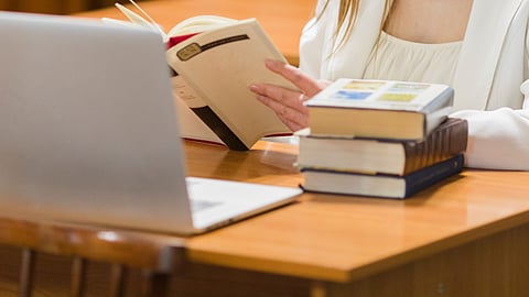 Image of books piled up over a wooden table beside a laptop and a lady in the background.
