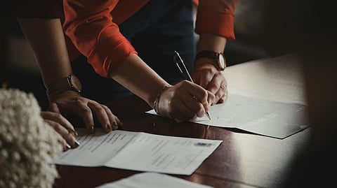 Image of 2 person one signing the document and other reading a paper.