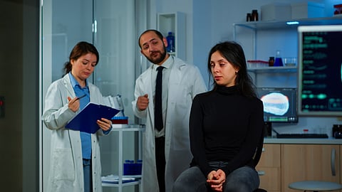 Woman waiting for doctor sitting on a chair wearing a black sweater and jeans, doctors are standing behind her.