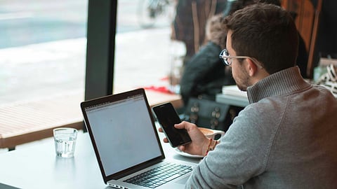 An image of a man sitting with his laptop and scrolling on phone.