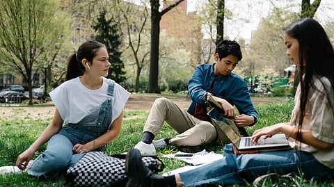 An image of 3 students with their laptops sitting in a park