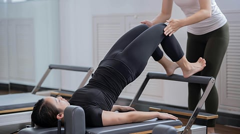 An image of a woman using Pilates equipment too exercise and another woman helping her.