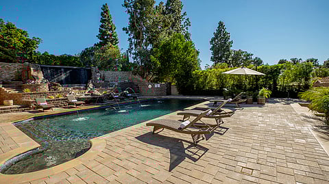 Image of a luxurious outdoor area with a swimming pool, patio, and lush greenery.