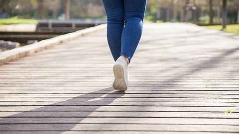 Woman wearing a blue jeans and white shoes walking on a wooden pathway in a park.